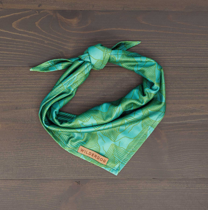 The Juniper Bandana, a green patterned quick-drying accessory with a “Wilderdog” label, is neatly tied and displayed on a dark wooden surface—perfect as a stylish cooling bandana for your dog.