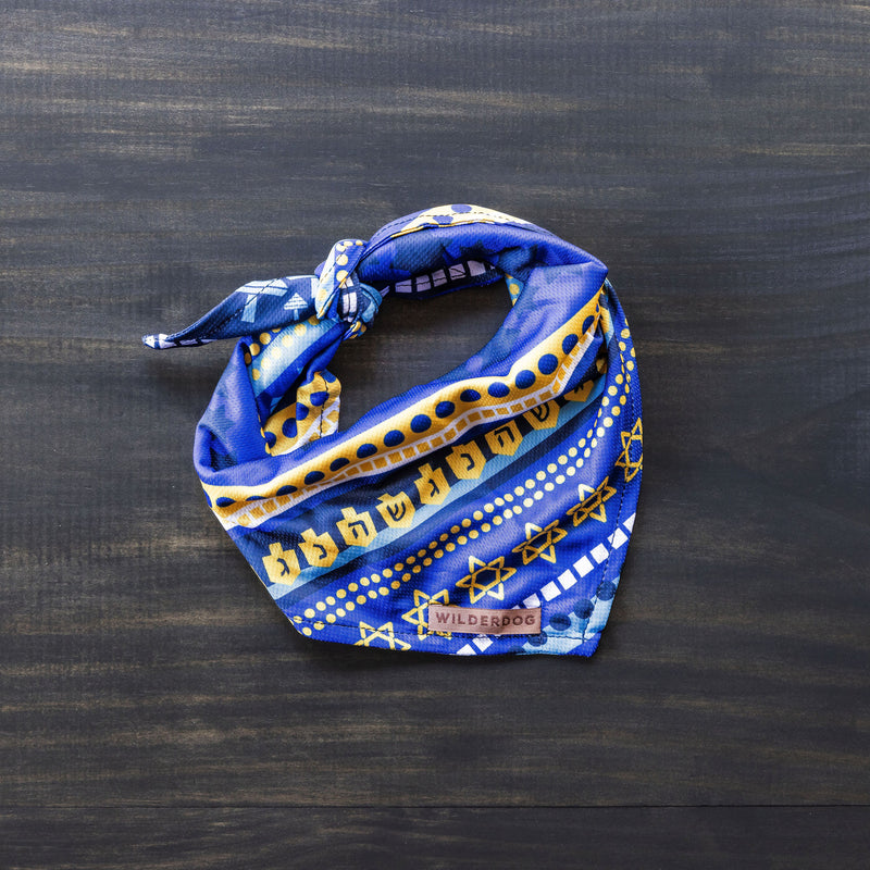 A vibrant blue and yellow Hanukkah Bandana featuring menorahs, dreidels, and Stars of David is neatly folded against a dark wooden background. Perfect for dog accessories, the "Wilderdog" brand logo is visible on the edge.