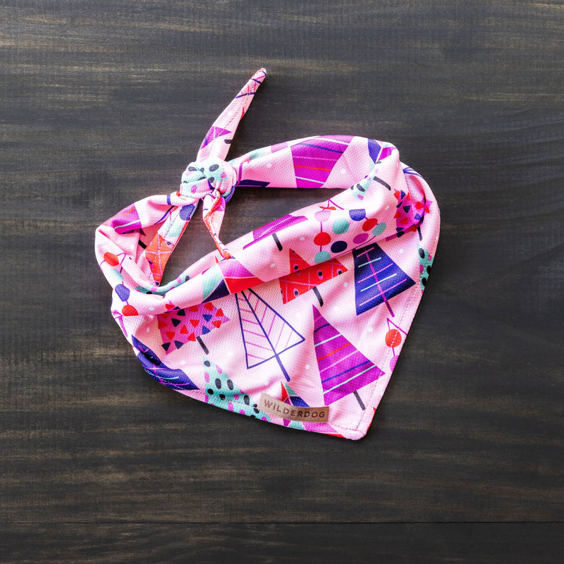 The Sugarplum Bandana, featuring a vibrant geometric tree pattern in shades of pink, purple, and red, lies flat on a dark wooden surface. Ideal for summer as pet gear, the cooling bandana is tied in a knot at one corner and has a small tag reading "Wilderdog.