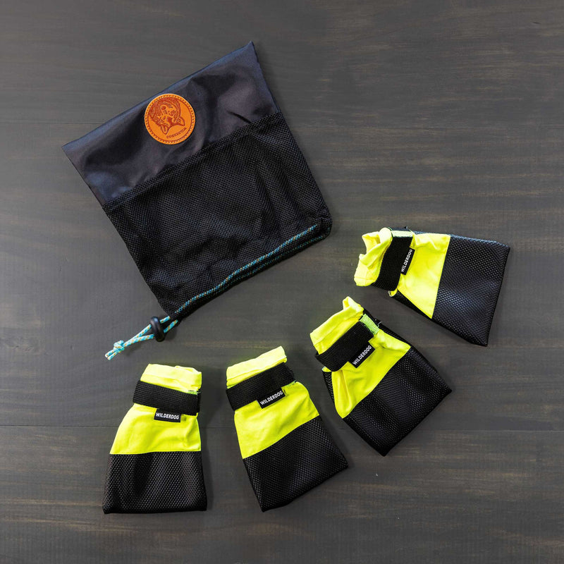 A set of Dog Booties, featuring four yellow and black booties with Velcro straps for excellent paw protection, is arranged in a semi-circle on a gray surface. Positioned above them is a black mesh storage bag with a drawstring and adorned with a brown leather patch.