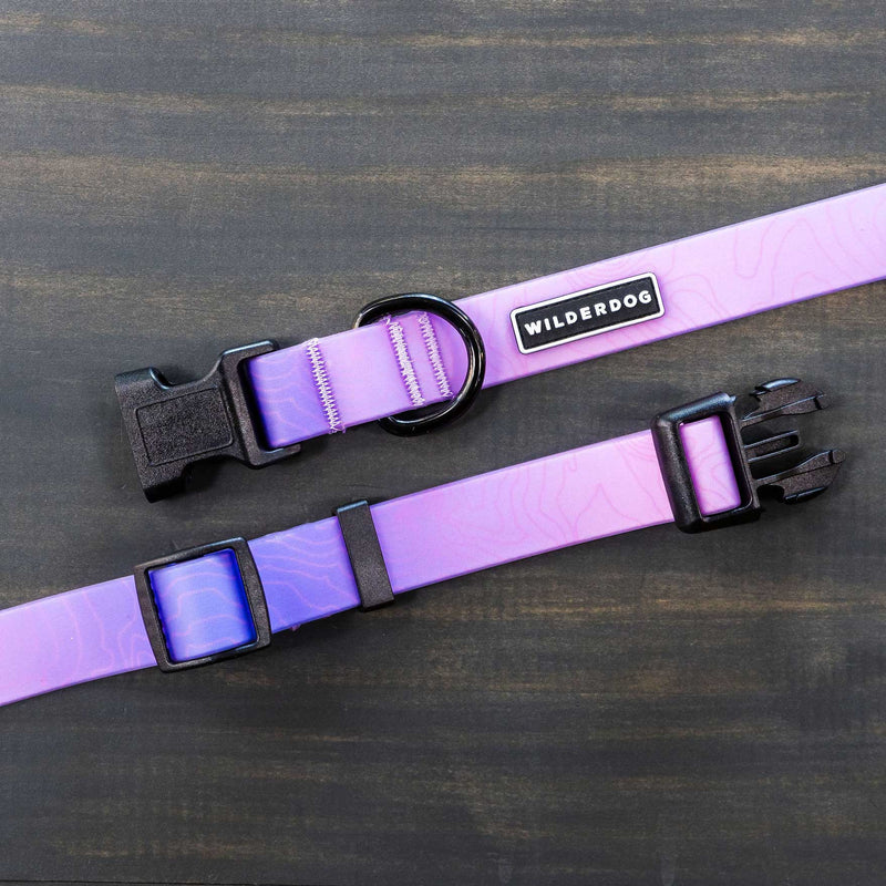 A close-up of the Aurora Waterproof Collar, a durable dog collar in purple featuring black hardware, a plastic buckle, adjustable strap, and a metal ring. The brand name "Wilderdog" is showcased on a small black and white label. The background is a dark wood surface.