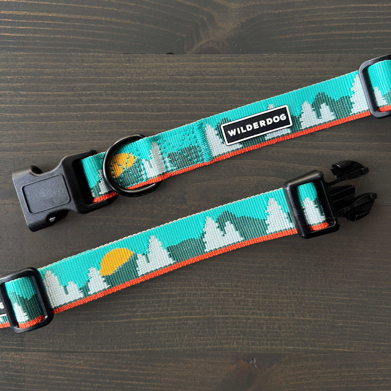 Two Tamarack Ultralight Collars, featuring turquoise nylon with mountain and sun designs, black plastic buckles, and "WILDERDOG" logo tags, are displayed on a dark wooden surface.