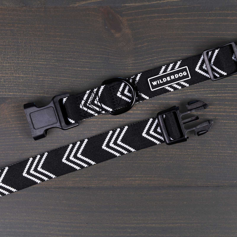 A black and white Orca Ultralight Collar with a zigzag pattern design is shown on a dark wooden surface. The collar features nylon webbing, a heavy-duty plastic clip, metal ring, and an adjustable strap. A tag on the collar reads "WILDERDOG.