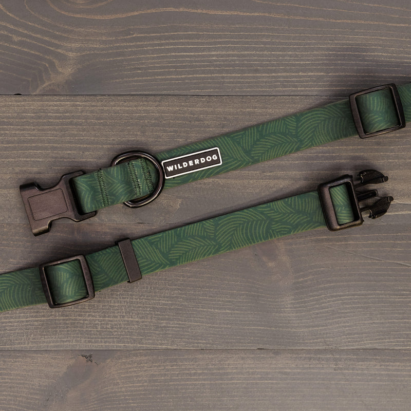 Two green Cedar Waterproof Collars with a leaf pattern and black hardware are laid out on wood. One collar features a "Wilderdog" label, metal D-ring for leash attachment, and is built for durability in all your adventures.