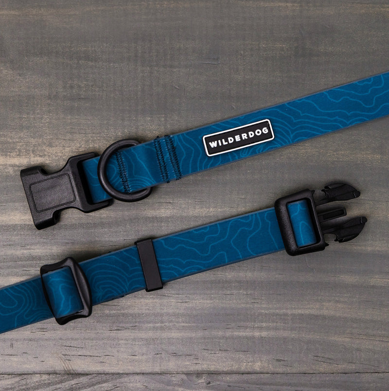 The Pacific Blue Waterproof Leash + Collar Bundle, featuring black plastic buckles, metal rings, a topographic pattern, and "WILDERDOG" label, rests on a gray wooden surface—an ideal addition to your durable dog gear collection.