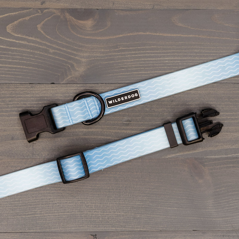 The Saltwater Waterproof Collar by Smartcart21 is a light blue, durable dog collar with white wavy lines, a black plastic buckle, adjustable slider, and metal ring, shown on a gray wooden surface.