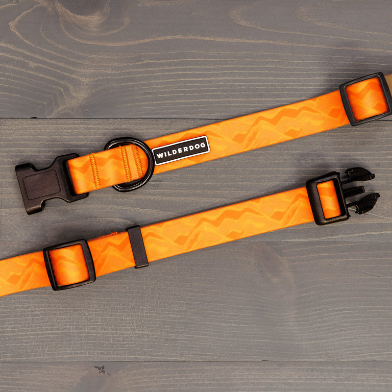 The Terracotta Waterproof Leash + Collar Bundle (x) features a zigzag-patterned orange collar with a black plastic buckle and "Wilderdog" tag, shown on gray wood—ideal for matching with durable dog gear.