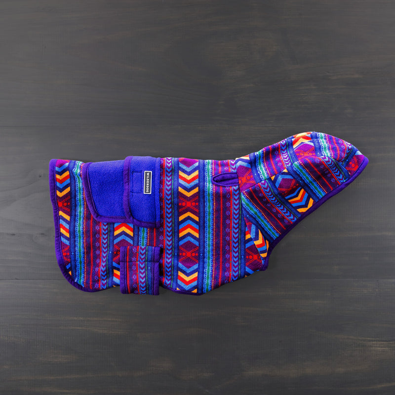 The Big Sky Fleece Jacket, designed by Smartcart21 and adorned with vibrant geometric patterns in blue, red, purple, and orange hues, is displayed flat on a dark wooden surface. This jacket features a hood and a Velcro strap for secure fastening. Enjoy peace of mind with our lifetime warranty.