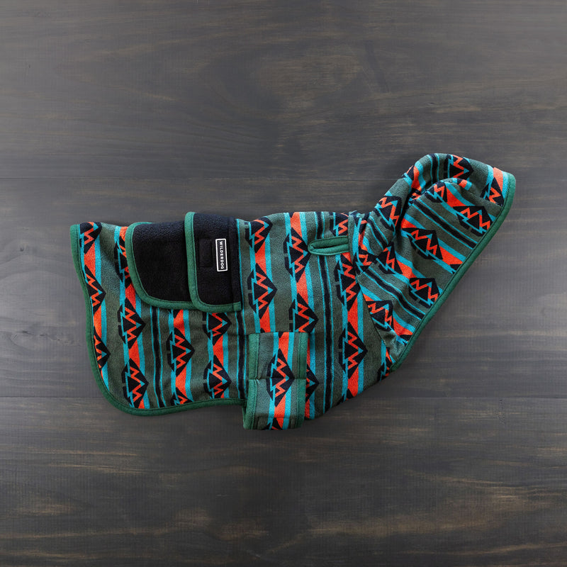 The Olive Fleece Jacket features a geometric pattern in teal, orange, red, and black. This stylish dog coat includes a black strap with Velcro for secure chest fastening and a hood. Laid flat on a dark wooden surface, the jacket's premium quality is highlighted, further supported by the Smartcart21 Lifetime Warranty.