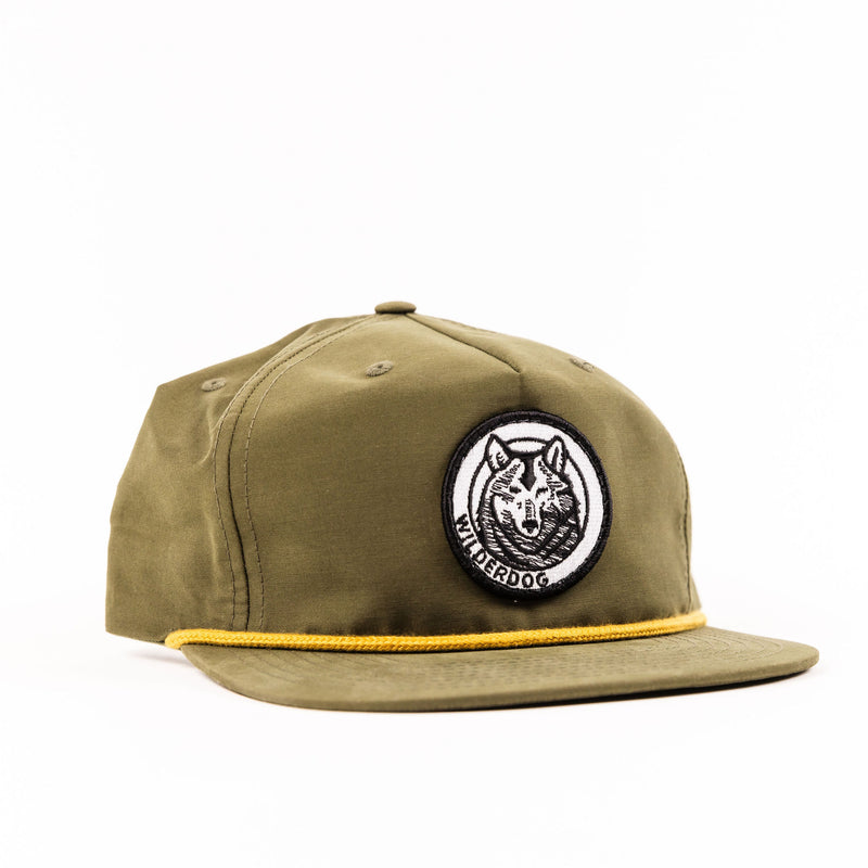 The Smartcart21 Gramps Hat is an olive green baseball cap with a flat brim and a circular embroidered patch on the front, featuring a stylized wolf head with the text "WILDERDOG" around it. Echoing classic golf vibes, this hat also includes a yellow rope detail across the base of the brim.
