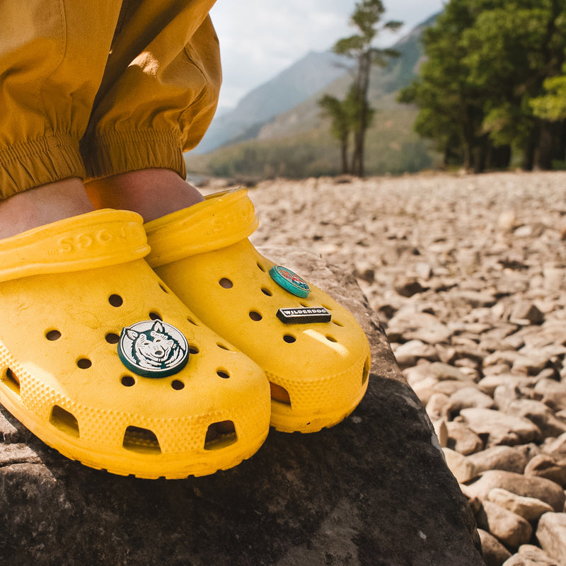 Close-up of a person's feet wearing bright yellow Crocs, decorated with Smartcart21 Shoe Charms featuring a rhino and the brand's logo, standing on a rocky surface against a backdrop of mountains and trees. The person is dressed in yellow pants.