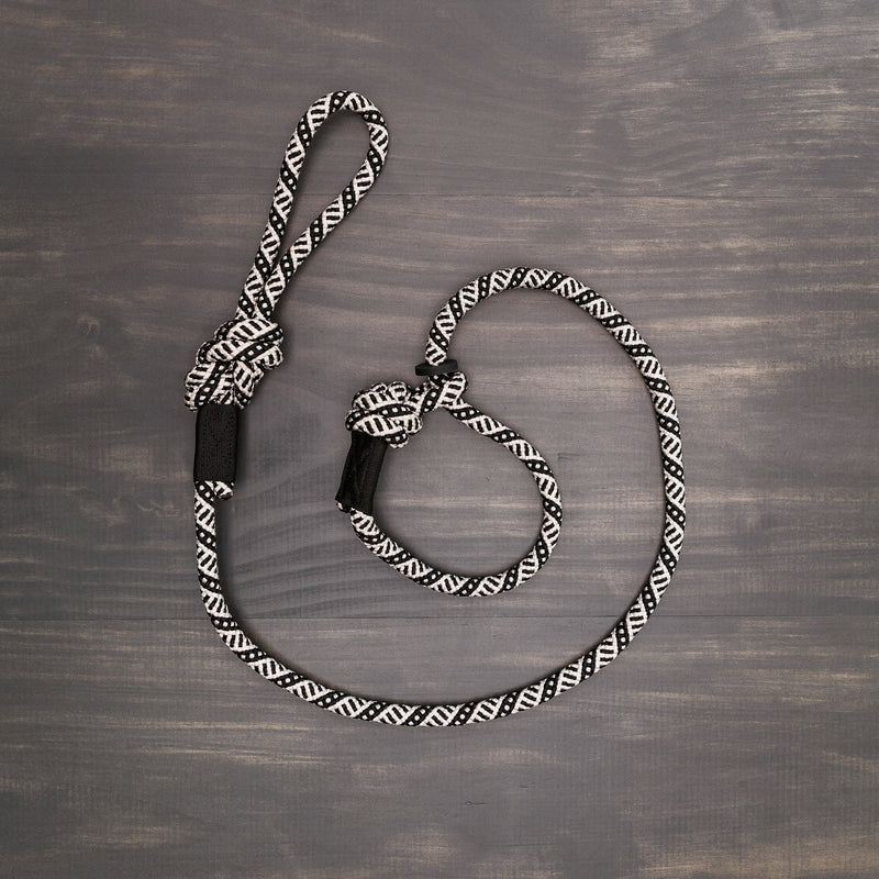 A coiled Black and White Slip Lead lies on a wooden surface. Made from durable rock climbing rope, it features a loop at one end and a knot near the other, likely designed for versatile use. The wood background has a dark, slightly weathered finish.