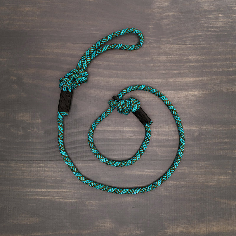 A Cascade Slip Lead in teal and black, resembling a rock climbing rope leash, is coiled neatly on a wooden surface. This durable lead features two knots: one larger knot near the middle and another smaller knot forming a loop at one end.
