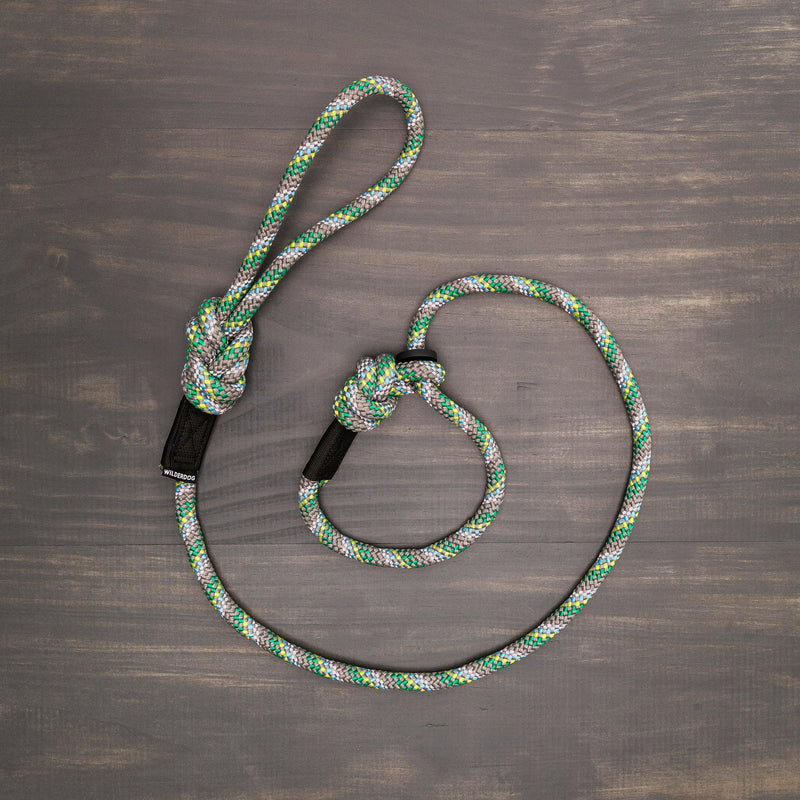 The Alpine Reflective Slip Lead, with its vibrant mix of green, white, and black colors, is displayed on a wooden surface. The durable rope is looped and twisted into a knot, resembling a rock climbing rope. The wooden background shows a greyish-brown tone with distinct grain patterns.