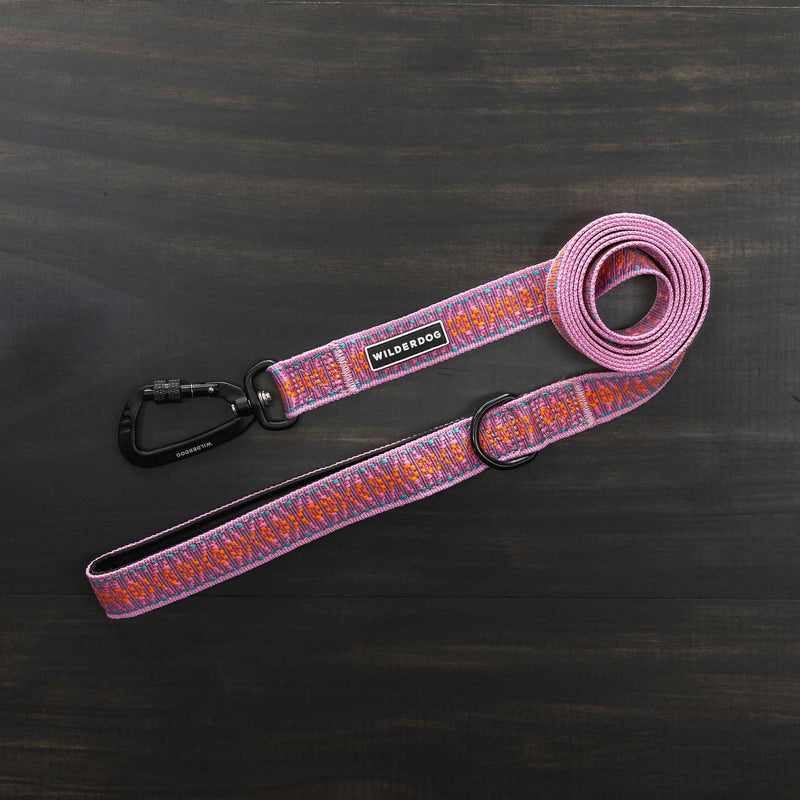 The Lilac Ultralight Leash, adorned with a vibrant pattern of pink, orange, and purple, is fully extended on a dark wooden surface. It features a black metal clasp and an adjustable padded handle. Part of the Ultralight Collection from Smartcart21, it includes a tag that reads "Wilderdog.