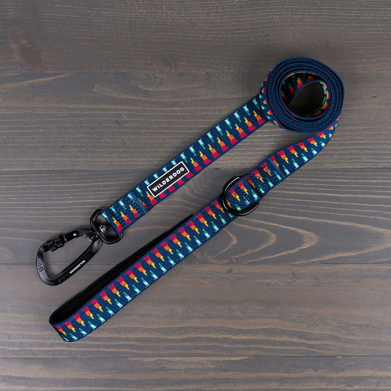 The Marina Ultralight Leash is a colorful dog leash with a blue background and multicolored geometric shapes, crafted from durable nylon webbing. It features a black swiveling carabiner clip and a looped handle. The brand name "Wilderdog" is visible on a label attached to the ultralight leash, which is placed on a dark wooden surface.