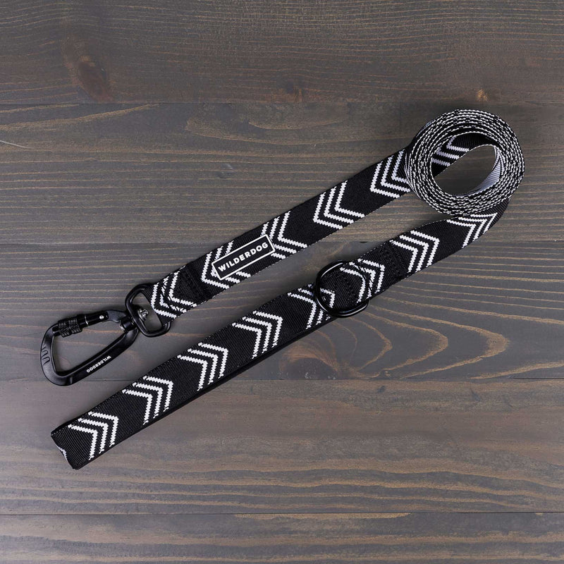 The Orca Ultralight Leash, featuring a black design with a white zigzag pattern, lies coiled against a dark wooden surface. A branded tag reading "Wilderdog" is prominently displayed on the lightweight nylon webbing. The ultralight leash includes a secure aluminum carabiner clip at the end.