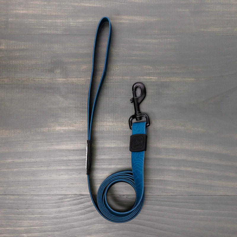 The Waterproof Leash & Collar Bundle (Pacific Blue) is coiled neatly on a gray wooden surface, featuring a durable blue design with a loop handle and black metal clasp.