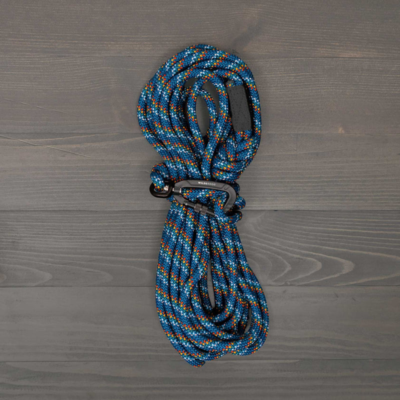 The Pacific Blue Reflective Long Lead - 30ft is a coiled blue, orange, and white rope leash with a black locking carabiner, shown on a gray wooden surface.