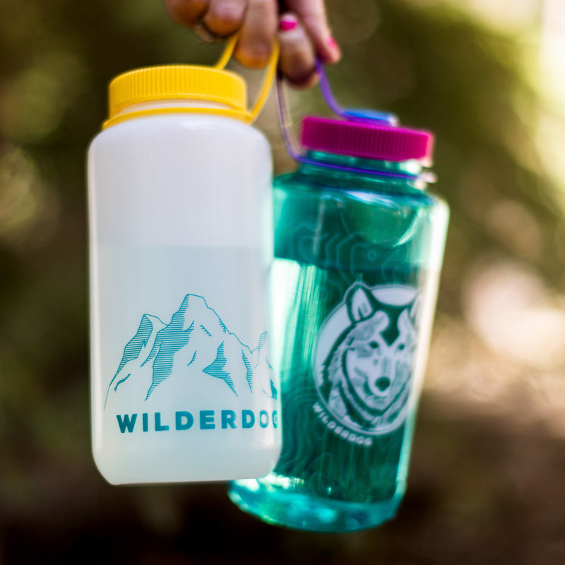 A person holding two colorful BPA/BPS-free Smartcart21 Nalgene water bottles; one is a 32oz Wide Mouth, white with a yellow cap, featuring a mountain graphic and the text "WILDERDOG." The other is teal with a purple cap, depicting a dog's face in a circle with the words "WILDERDOG" around it.