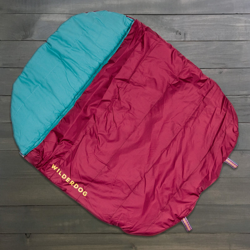 A maroon and teal Sleeping Bag with "WILDERDOG" in yellow on one side, laid flat on dark wood. Crafted from durable ripstop polyester, it's ideal for outdoor adventures with your furry friend.