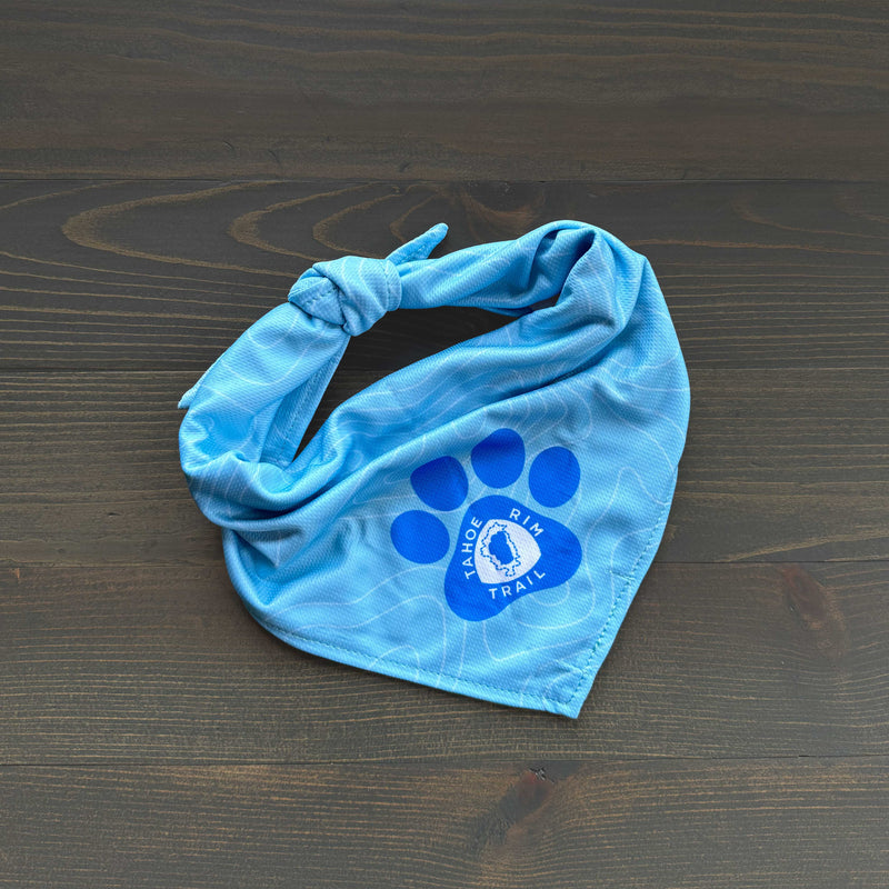 A Tahoe Rim Trail Association Bandana in light blue, tied with a knot at one end, rests on a dark wooden surface. It showcases a dark blue paw print design with the text "Tahoe Rim Trail" surrounding an image of a trail hiker in the center, making it an ideal pet accessory for adventurous outings.