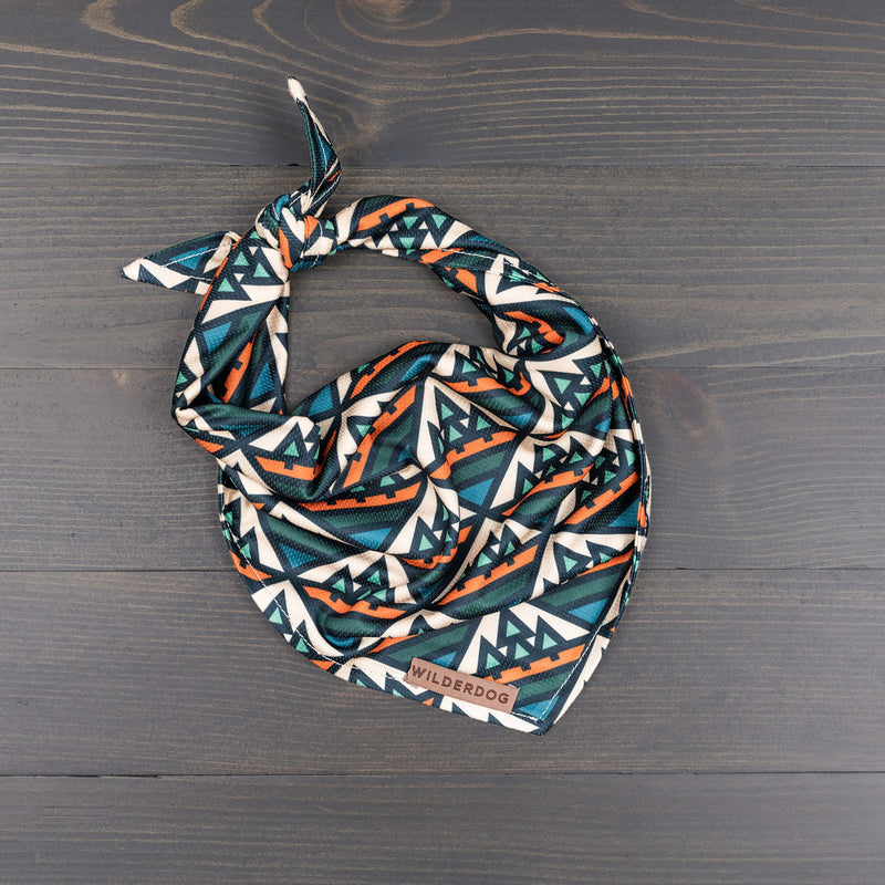 Camp Bandana with teal, orange, and white triangles, twisted and knotted on a dark wooden surface.