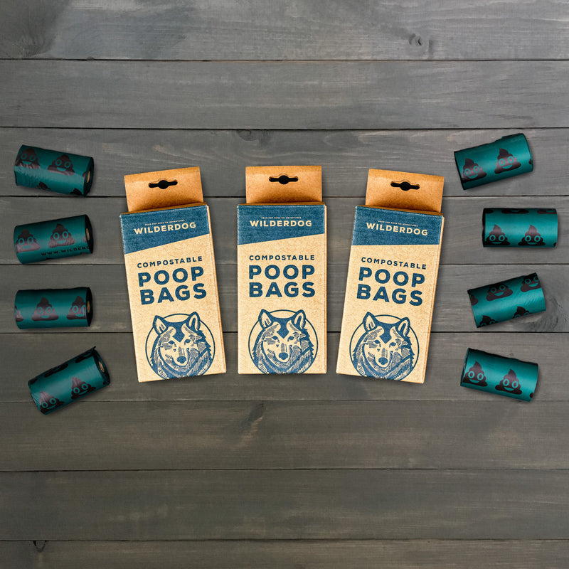 Three boxes of Compostable Dog Poop Bags - 3 Pack are centered, each showcasing an illustration of a dog's face. These are encircled by seven rolls of dark green biodegradable bags decorated with paw print designs, all thoughtfully displayed on a wooden surface.