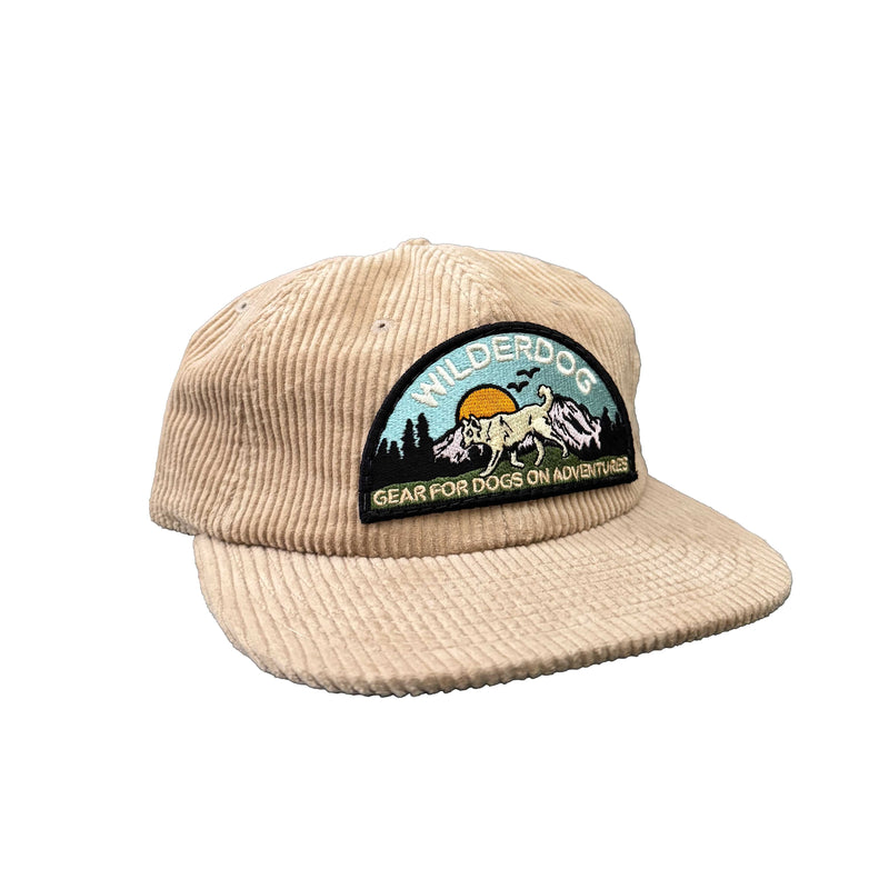 The Smartcart21 Cord Hat is a beige 100% cotton cap with a vibrant dog, sunburst, and trees patch. It features "Wilderdog" and "Gear for Dogs on Adventures" embroidery, plus an adjustable clasp for the perfect fit.