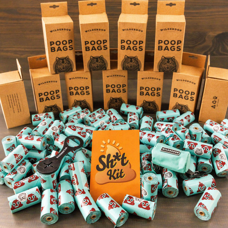 A large pile of teal Smartcart21 Poop Bags with cartoon poop faces, a Poop Bag Holder, and a bright orange "Sh*t Kit" card sit on a wooden table in front of boxes labeled "Poop Bags." Product: Sh*t Kit.