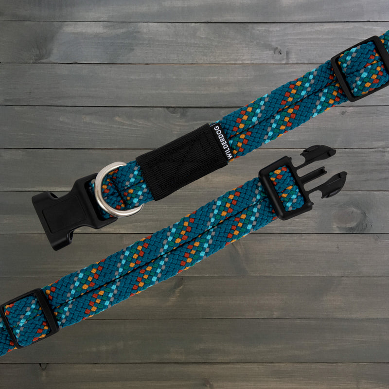 Two Pacific Blue Reflective Collars, known for their durability and eye-catching geometric pattern, are laid out on a wooden surface. Inspired by rock climbing rope collar designs, these teal-colored collars feature black plastic buckles, metal D-rings, and a fabric tag that reads "Wilderdog.