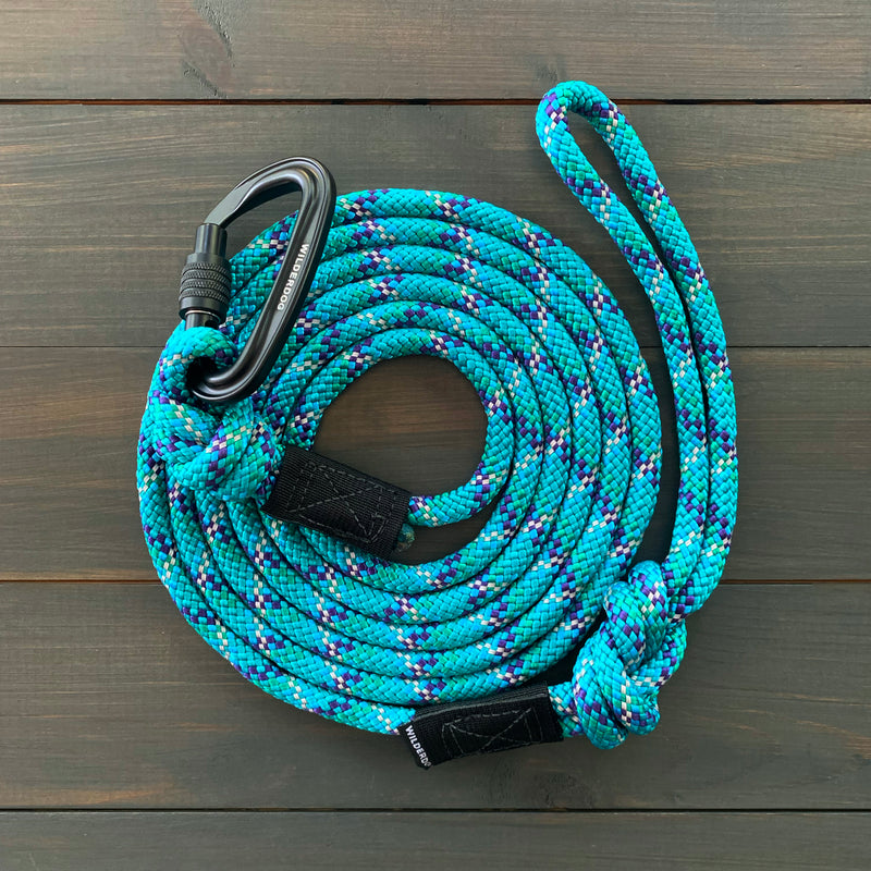 A coiled Islander Reflective Leash in blue and turquoise, adorned with purple and white patterns, features a black carabiner attached at one end. It lies on a dark wooden surface, with one end of the leash looped and the other end knotted.
