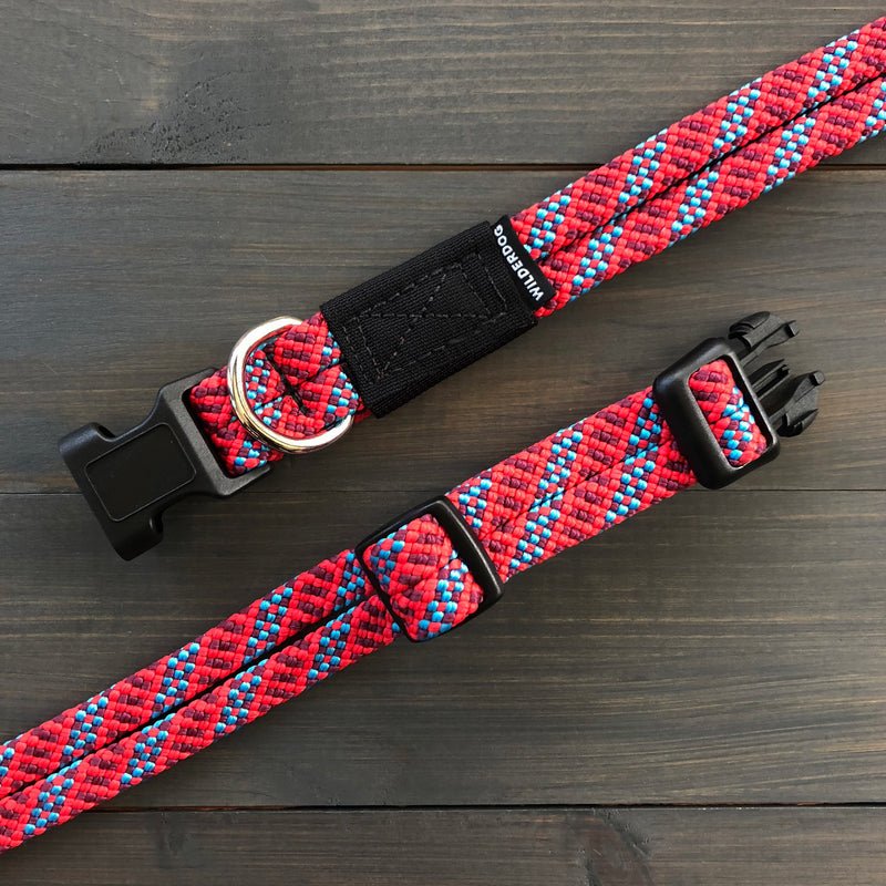 A stylish Maple Collar, made from durable rock climbing rope in red and blue colors, featuring black plastic buckles and adjustable fittings, is displayed on a dark wooden surface. The collar includes a silver D-ring for attaching a leash and a small black fabric label with white text.