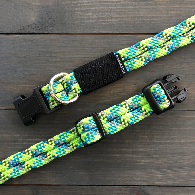 The Lime Reflective Collar, a vibrant multicolored dog collar featuring a mix of green, blue, and yellow hues, is displayed on a dark wooden surface. The collar boasts heavy-duty hardware, including a black plastic buckle, a metal D-ring, an adjustable length with a black tag that reads "WILDROO.