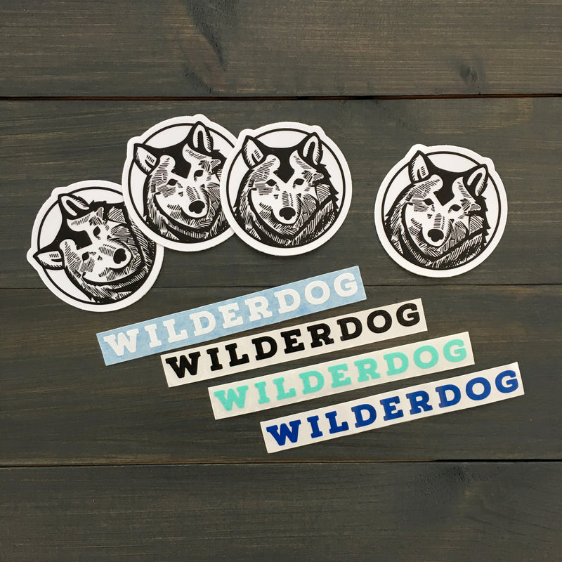 An image displays four circular stickers with a stylized dog's face, accompanied by five Smartcart21 Sticker Decals in various colors and fonts. These outdoor-rated, waterproof vinyl decals are neatly arranged on a dark wooden surface.