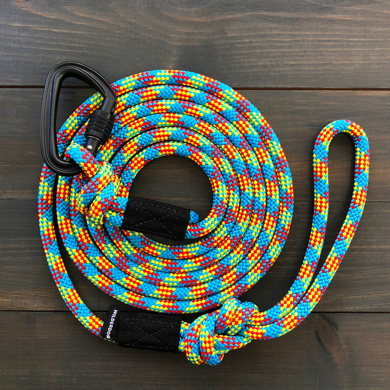 An Aprés Leash coiled neatly with a durable carabiner mechanism attached to one end and a loop at the other, displayed on a wooden surface. The leash features a vibrant pattern of blue, yellow, red, and white.
