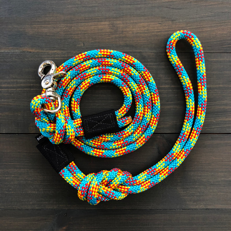 The Aprés Quick Clip Leash, an intricately braided climbing rope leash adorned with a vibrant mix of blue, orange, and yellow colors, features a quick clip clasp. Carefully coiled on a dark wooden surface, this durable dog leash includes a woven handle and a knot near the clasp end.