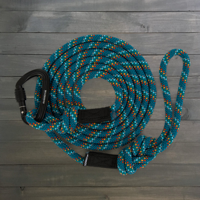 A coiled blue and orange item with a carabiner attached on the left end, the durable Pacific Blue Reflective Leash is neatly arranged on a gray wooden surface. The leash features black accent bands securing the loops, ensuring reliability and strength for all your outdoor adventures.