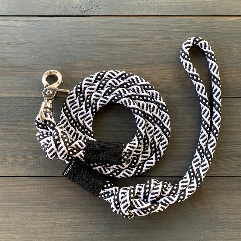 The Black and White Quick Clip Leash, featuring a durable woven design reminiscent of rock climbing ropes, is coiled on a wooden surface. Its black and white pattern is complemented by a metallic quick clip for attaching to a collar at one end, and it includes a handle loop at the other end.
