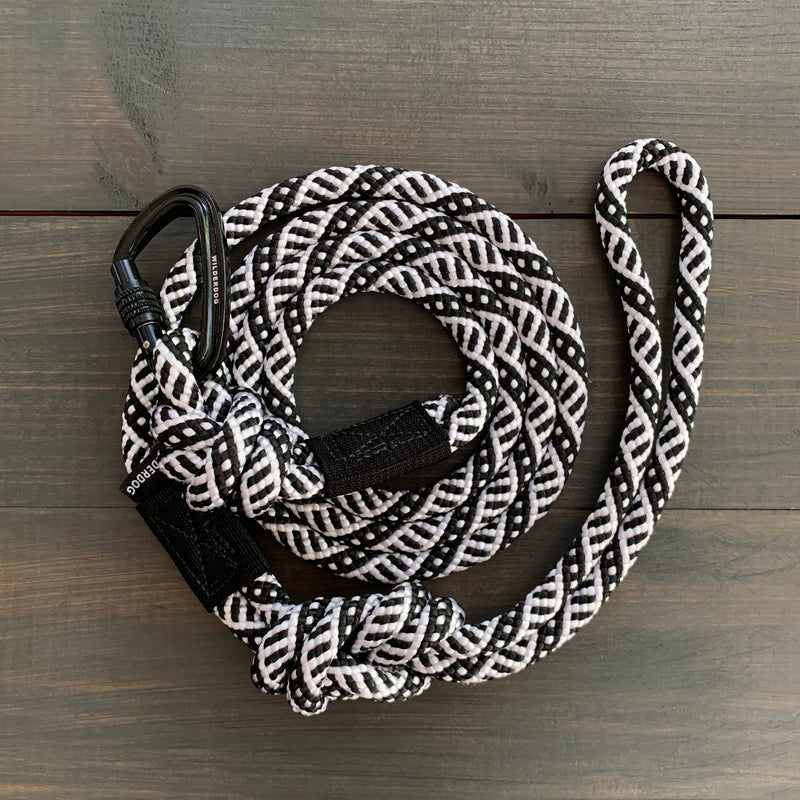 A Black and White Small Carabiner Leash, featuring a checkered pattern of intertwined black and white strands, is coiled on a wooden surface. One end of the leash has an attached carabiner lock.