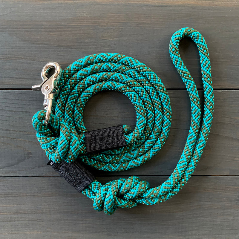 The Cascade Quick Clip Leash, a durable green and black coiled rock climbing rope leash with a silver metal clasp, is neatly arranged on a wooden surface. It features a loop handle and a black fabric strap near the clasp end for added grip.
