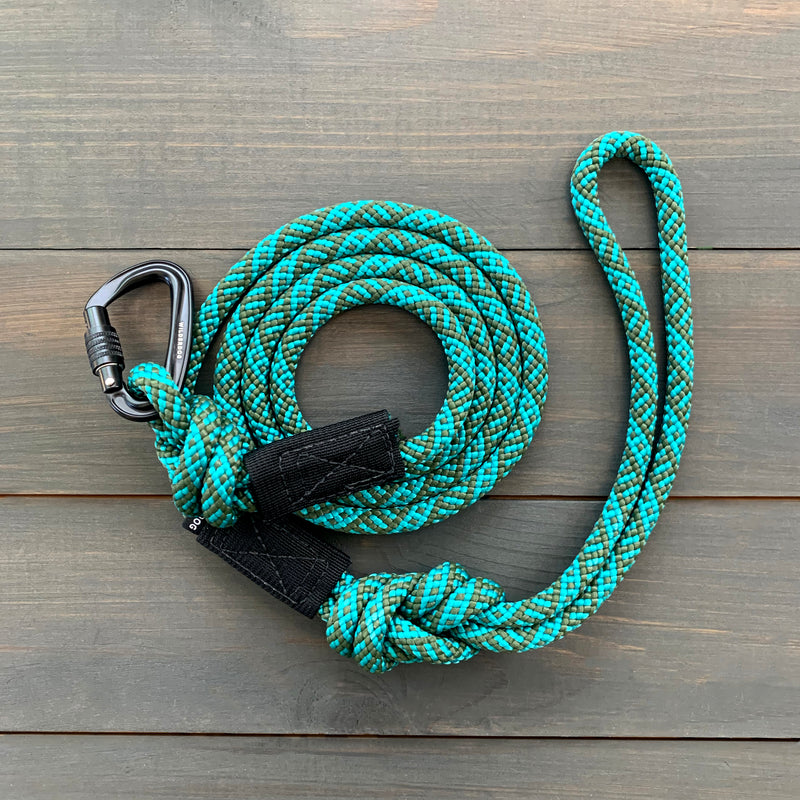 A coiled turquoise and black Cascade Small Carabiner Leash, renowned for its durability, rests on a wooden surface with a carabiner attached to one end. The carabiner and the ends of the leash are reinforced with black fabric. This versatile tool also functions as a durable climbing rope and comes with a lifetime warranty.