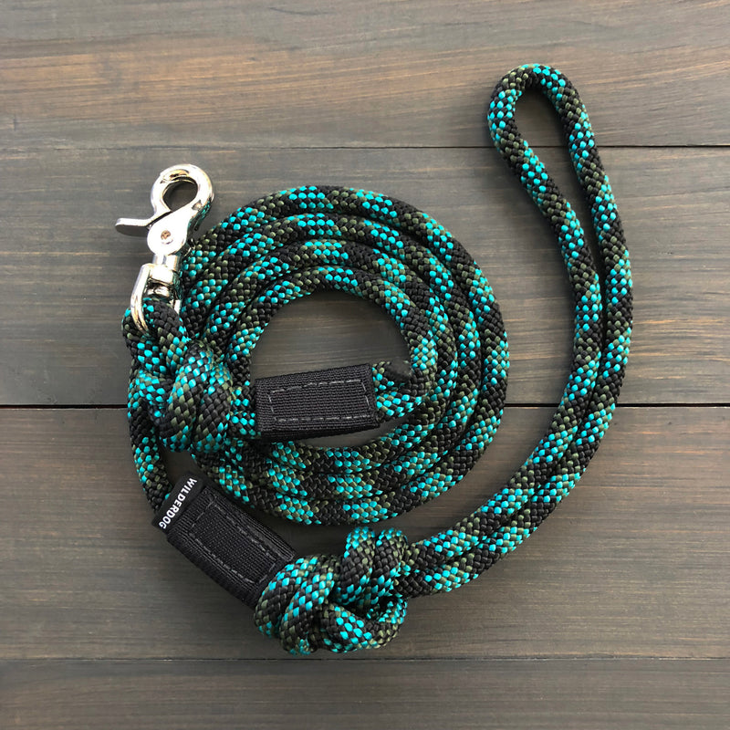 A Granite Quick Clip Leash, featuring a coiled black and teal braided pattern resembling durable rock climbing rope and a silver metal clasp, is laid on a wooden surface. This leash includes a loop handle at one end and a quick clip clasp at the other for attaching to a collar or harness.