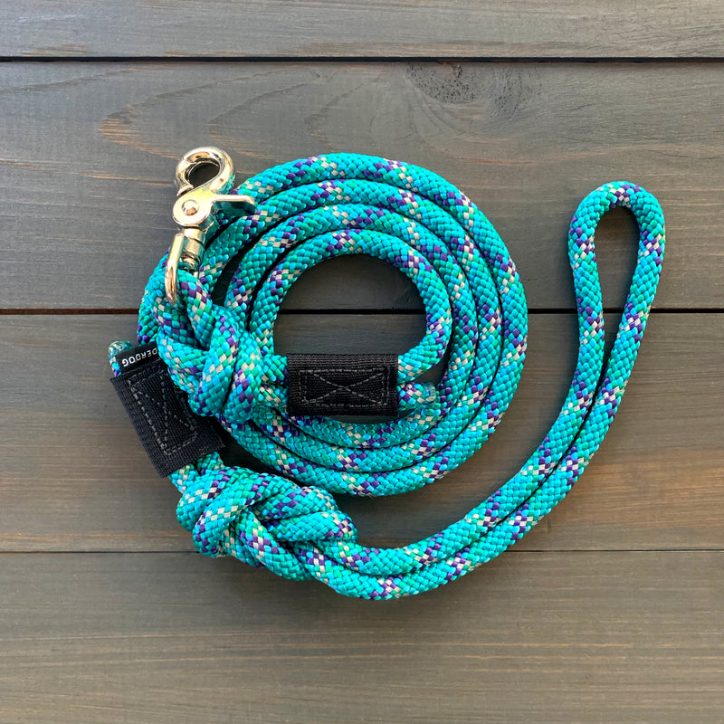 The Islander Reflective Quick Clip Leash, made of durable braided rope, is neatly coiled on a wooden surface. Predominantly teal with interwoven patterns of purple, white, and blue, it features a sturdy metallic clip and a black fabric loop at the handle for a secure grip. This rock climbing rope leash ensures strength and reliability.