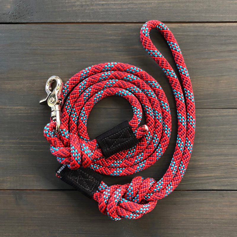 The Maple Quick Clip Leash, featuring a coiled design with a red, blue, and white pattern and a metal clip, lies on a dark wooden surface. The leash includes a wrist loop at one end and a quick clip for attaching to a collar at the other end.