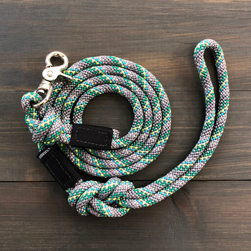 The Alpine Reflective Quick Clip Leash, a coiled multicolored rope dog leash made from durable rock climbing rope with a metal clasp, is neatly arranged on a dark wooden surface. The pattern on the leash includes shades of gray, green, and yellow, and a loop handle is visible at one end. Small black fabric labels are attached near the clasp and handle.
