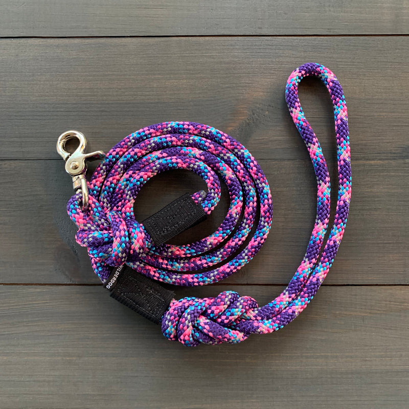 The Razzleberry Reflective Quick Clip Leash, a colorful and durable braided dog leash with a silver metal clasp, is coiled neatly on a dark wooden surface. This leash showcases vibrant patterns in purple, blue, pink, and white, with its prominently visible handle loop adding to its appealing design.