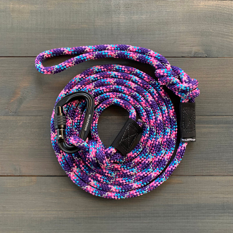The Razzleberry Reflective Leash is a coiled, multi-colored, durable dog leash equipped with a black carabiner clip at one end. The leash boasts an intricate pattern of purple, pink, blue, and white. It's displayed on a dark, wooden surface that highlights its vibrant colors and sturdy design similar to rock climbing rope leashes.