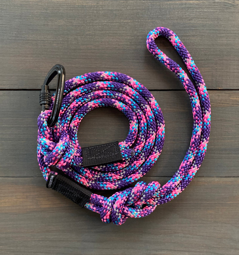 The Razzleberry Reflective Small Carabiner Leash, coiled on a wooden surface, showcases a vibrant mix of pink, blue, purple, and white colors woven together from durable rock climbing rope. It features a loop handle at one end and a sturdy black carabiner lock for secure attachment.