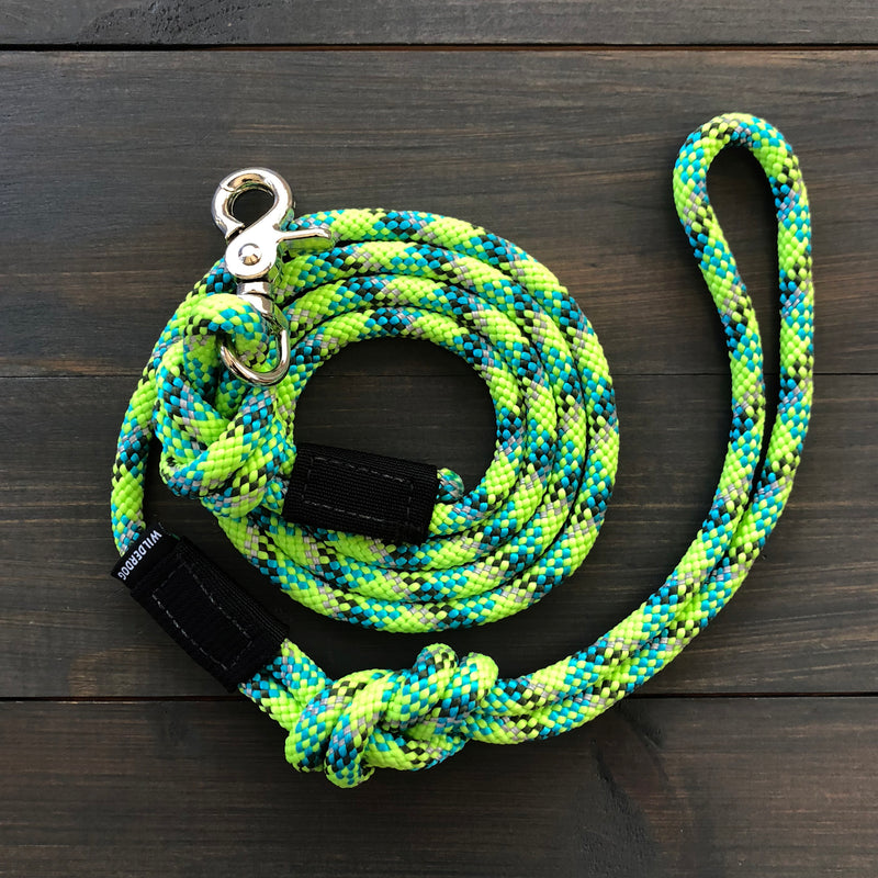 The Lime Reflective Quick Clip Leash is a coiled, multicolored durable dog leash with shades of green, blue, and yellow. It features a loop handle and a silver metal clasp for attaching to a collar. Made from rock climbing rope, it is laid out on a dark wooden surface.
